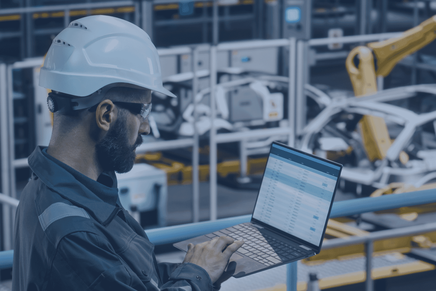 Engineer in hard hat using a laptop on a factory floor with robotic manufacturing equipment behind