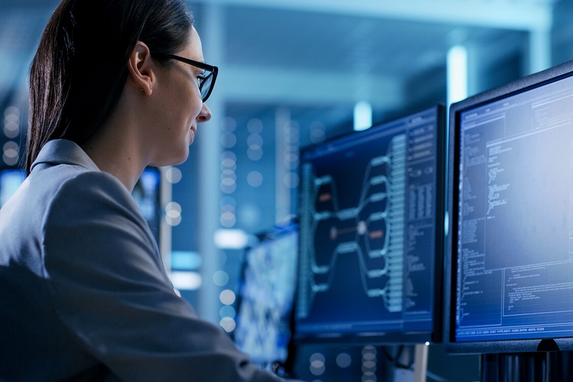 Engineer at workstation with multiple monitors showing data-flow diagrams and code in a dark blue server room